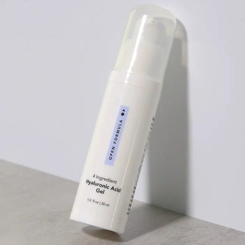A white pump bottle of Open Formula Hyaluronic Acid Gel, designed to hydrate dry skin and reduce lines and wrinkles, stands upright on a gray surface with a light background. The label displays black text.