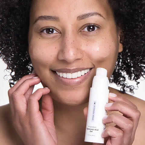 A smiling person with curly hair applies Open Formula Hyaluronic Acid Gel to their cheek, holding a white bottle labeled “Hyaluronic Acid Gel.” Their radiant, healthy skin appears moisturized and free of dryness.