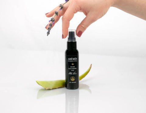 A hand with painted nails holding lavender hovers above a black AREMES FERMENTIS Aloe Vera Hand Cleansing Gel bottle, offering hydrating and sanitizing benefits, next to an aloe vera leaf on a white background.