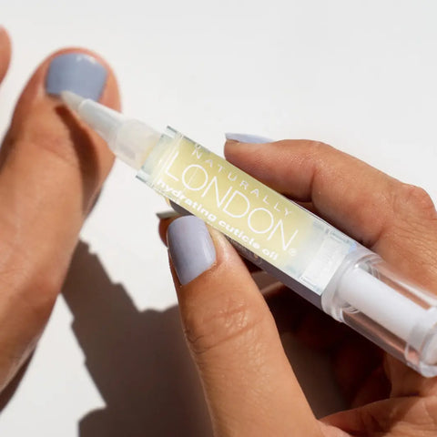 A hand uses the Naturally London Hydrating Cuticle Oil, 9mL—a botanical nail care oil—in a pen-style applicator on light blue polished nails, set against a white background.