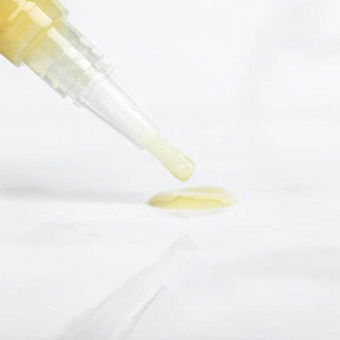 A close-up of a dropper releasing a small drop of Naturally London Hydrating Cuticle Oil, 9mL, onto a white surface, with the oil and dropper faintly reflected below.
