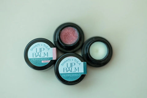 Three small black containers of Shanti + Tula Hydrating Natural Lip Balm sit on a light surface; one holds pink-tinted balm, another white, and the third displays the label—all offer lightweight, non-greasy hydration.