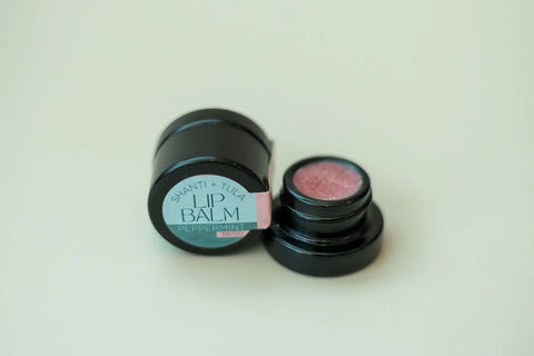 A small black container of Shanti + Tula Hydrating Natural Lip Balm – Non-Greasy, Lightweight & USA-Made Moisture Stick sits open on a light-colored surface, revealing the pink balm inside.
