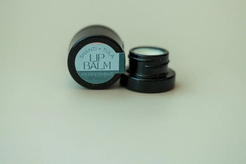 A black jar of Shanti + Tula Hydrating Natural Lip Balm – Non-Greasy, Lightweight & USA-Made Moisture Stick in peppermint flavor sits with its lid off, revealing the balm inside against a plain light background.
