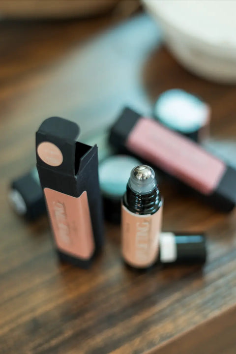 A close-up of the Shanti + Tula Hydrating Natural Lip Oil with its rollerball applicator exposed, shown alongside its packaging and similar clean beauty products on a wooden surface. Made in Utah.