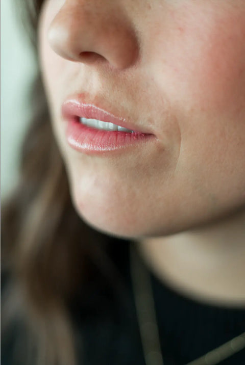 Close-up of smooth skin and slightly parted lips wearing light pink gloss from Shanti + Tulas Hydrating Natural Lip Oil – Rollerball Applicator. Clean beauty, made in Utah. Background and clothing are softly blurred.