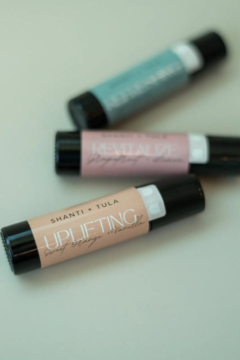 Three Shanti + Tula Hydrating Natural Lip Oil bottles with rollerball applicators lie diagonally on a light surface. Labeled UPLIFTING, REVITALIZE, and DEEP CALM, each is made in Utah with clean, natural ingredients.