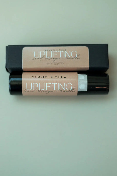 A tube of Shanti + Tula Hydrating Natural Lip Oil – Rollerball Applicator, a clean beauty product made in Utah, sits on a light surface with its black box packaging behind it.