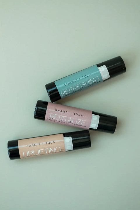 Three Hydrating Natural Lip Oil tubes from Shanti + Tula, in teal, pink, and peach, are labeled “REFRESHING,” “REVITALIZE,” and “UPLIFTING.” Each clean beauty rollerball rests on beige with a black cap. Made in Utah.