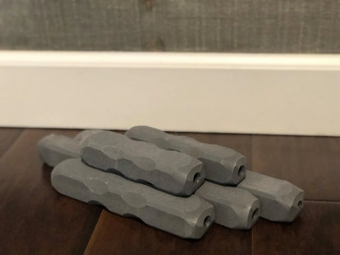 Five Indestructibone Ultimate Kennel Pack chew toys by Bulletproof Pet Products Inc—five gray, textured cylinders with hexagonal ends and central holes—are stacked on a dark wooden floor, perfect for tough chewers.
