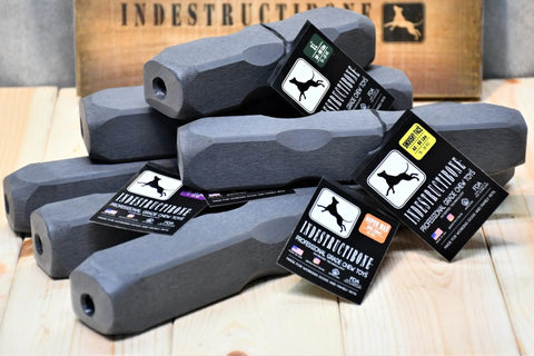 Five Indestructibone Super Ultimate Kennel Pack professional-grade chew toys by Bulletproof Pet Products Inc are stacked, each durable, bone-shaped and dark gray with a dog tag. A wooden box is in the background. Available in 6 sizes.