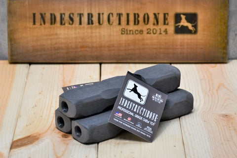 Three black Indestructibone Professional Grade Mini chew toys (3 Pack) for dogs up to 15 lbs. by Bulletproof Pet Products Inc are stacked on wood, with a INDESTRUCTIBONE Since 2014 sign and a dog logo in the background.