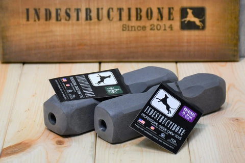 Two gray, bone-shaped Indestructibone™ Professional Grade toys (Original and XL Combo Pack) by Bulletproof Pet Products Inc rest on wood, with a sign behind them reading “INDESTRUCTIBONE Since 2014” and a dog silhouette logo.