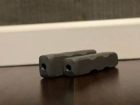 Two gray Indestructibone™ Professional Grade chew toys (Original and XL Combo Pack) by Bulletproof Pet Products Inc are on a wooden floor near a white baseboard and textured wall.