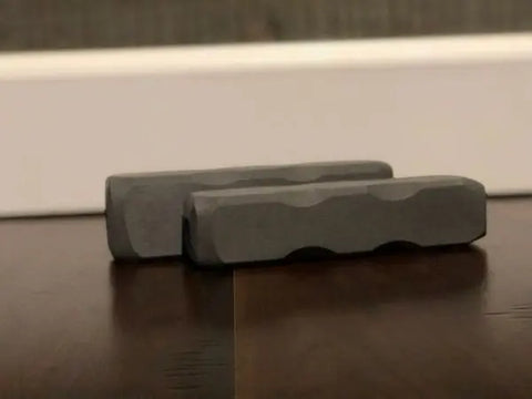 Two rectangular, gray Indestructibone™ chews by Bulletproof Pet Products Inc—Original and XL from the Professional Grade Combo Pack—rest side by side on a dark wooden surface in front of a white baseboard and textured wall.