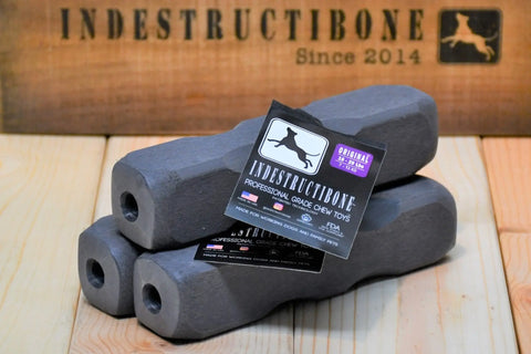 Three gray Indestructibone™ Professional Grade Original chew toys (3 pack, for dogs 16-29 lbs.) by Bulletproof Pet Products Inc are stacked on a wooden surface near a branded box featuring the logo and INDESTRUCTIBONE Since 2014.