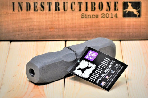 A gray Indestructibone™ Professional Grade Original chew toy for 16–29lb dogs by Bulletproof Pet Products Inc sits on wood, product tag attached. Behind it, a box shows the Indestructibone logo and Since 2014—ideal for tough chewers.