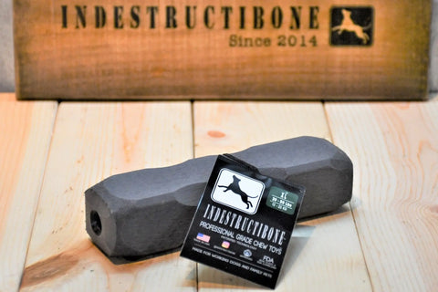 A black, hexagonal Indestructibone™ Professional Grade XL by Bulletproof Pet Products Inc for dogs 30-50 lbs sits on a wooden surface with its tag; behind it is a sign reading INDESTRUCTIBONE Since 2014 with a dog logo.