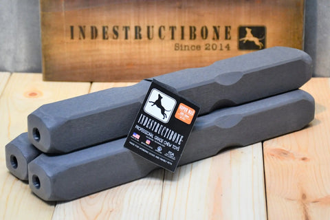 Three Indestructibone Professional Grade Super Max chew toys (3-pack) by Bulletproof Pet Products Inc, made for dogs 101 lbs+, are stacked on wood. A wooden sign behind reads INDESTRUCTIBONE Since 2014 with a dog silhouette.