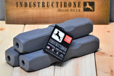 Three gray Indestructibone Professional Grade XL Plus chew toys for dogs 51-100 lbs are stacked on a wooden surface. A label and a Bulletproof Pet Products Inc sign highlight this durable 3-pack in the background.