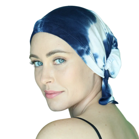 A woman with light skin and blue eyes wears the Elise Chemo Beanie® by Chemo Beanies®, featuring a blue and white tie-dye design. She looks over her shoulder with a soft smile, dressed in a sleeveless top against a plain background.