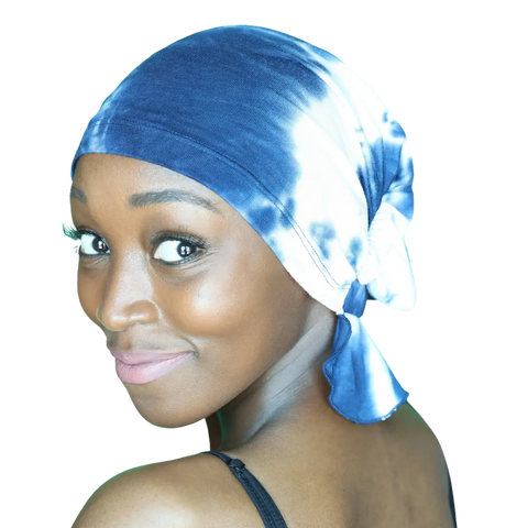 A woman with glowing brown skin smiles over her shoulder, wearing the Elise Chemo Beanie® by Chemo Beanies®, featuring blue and white tie-dye, paired with a black strap top against a white background.
