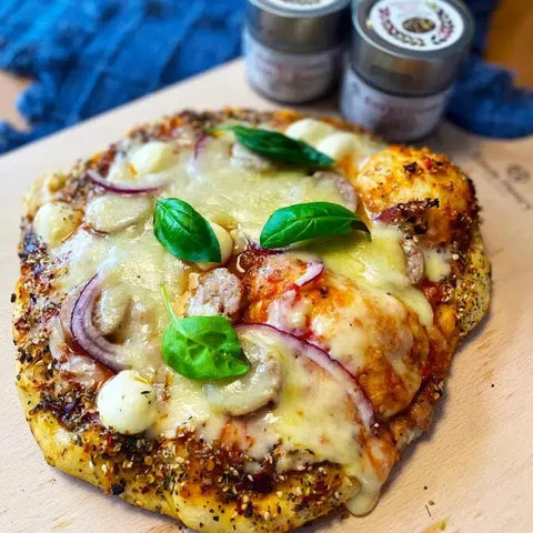 A homemade pizza with cheese, red onion, mushrooms, mozzarella balls, herbs, and basil rests on a wooden board. In the background are two blurred jars from the Gustus Vitae Italian Seasoning Gift Set – Tastes of Italy artisanal spice blends.