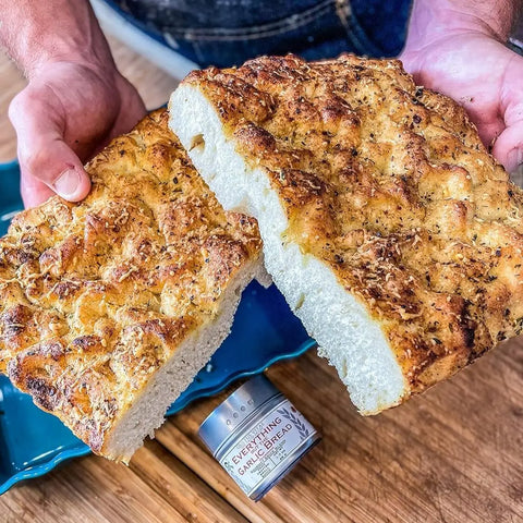 A person holds two halves of herbed focaccia. On the wooden table below rests a Gustus Vitae Italian Seasoning Gift Set – Tastes of Italy: an artisanal spice blends six pack.
