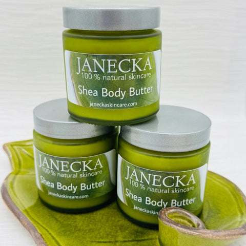 Three green jars of JANECKA Shea Body Butter by Janecka Skincare are stacked on a green leaf-shaped dish. Each jar reads 100% natural skincare and has a silver lid, shown against a white background.