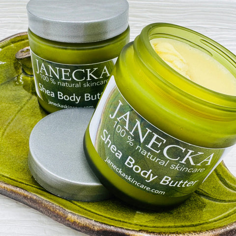 Two green jars of JANECKA Shea Body Butter by Janecka Skincare sit on a green dish—one open, showing the creamy moisturizer. Both are labeled as 100% natural skincare, ideal for eczema and psoriasis.