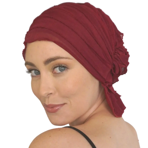 A woman with light skin, blue eyes, and light makeup wears the JEWEL CHEMOBEANIES® by Chemo Beanies®, looking over her shoulder and smiling slightly against a plain white background.