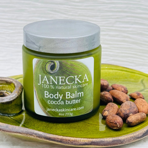 A green jar of JANECKA Body Balm, Cocoa Butter from Janecka Skincare sits on a green ceramic tray beside cocoa beans. The label highlights it as 100% natural and waterless skincare.