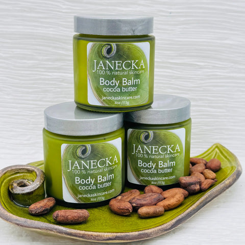 Three green jars of JANECKA Body Balm, Cocoa Butter by Janecka Skincare are stacked on a ceramic dish and surrounded by cocoa beans. Their labels read “100% natural skincare” against a light, minimal background.
