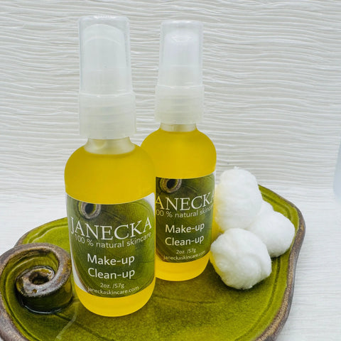 Two bottles of JANECKA Make-Up Clean-Up by Janecka Skincare, a natural eye makeup remover, sit on a green ceramic dish with three cotton balls beside them, all set against a white background.