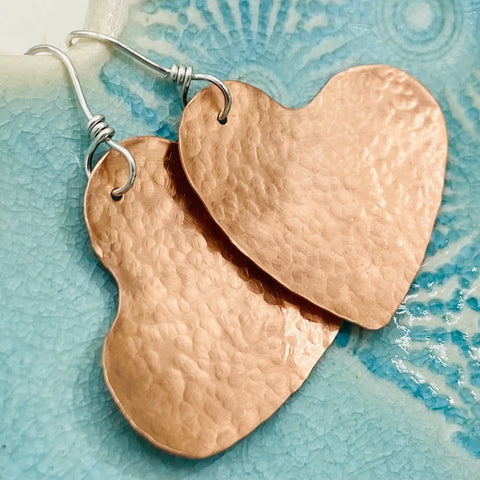 Janecka’s JANECKA Copper Hand Forged Heart Earrings with sterling silver ear wires are displayed on a textured blue ceramic surface featuring a seashell pattern.
