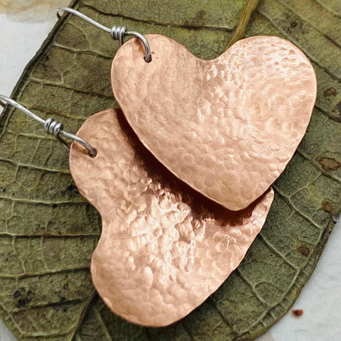 JANECKA Copper Hand Forged Heart Earrings by Janecka feature sterling silver ear wires and are displayed on a dried leaf—an elegant piece of handmade jewelry.