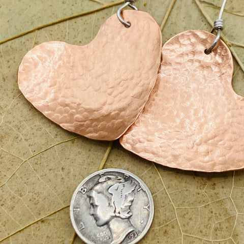 Close-up of JANECKA Copper Hand Forged Heart Earrings by Janecka, featuring large copper hearts with sterling silver ear wires, shown beside a 1936 Mercury dime for scale on a textured leaf backdrop—a beautiful handmade jewelry piece.