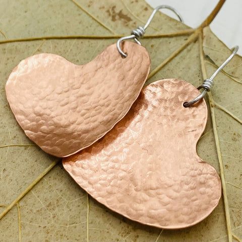 The Janecka JANECKA Copper Hand Forged Heart Earrings feature sterling silver ear wires and a textured, handcrafted look, displayed on a dried leaf background for unique appeal.