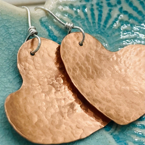 Close-up of JANECKA Copper Hand Forged Heart Earrings by Janecka, featuring sterling silver ear wires and displayed on a blue ceramic surface with a subtle shell-like texture.