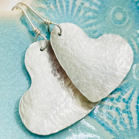 A close-up of Janecka’s JANECKA Sterling Silver Hand Forged Heart 1.5 Earrings, handcrafted and heart-shaped, displayed on a textured light blue ceramic surface.