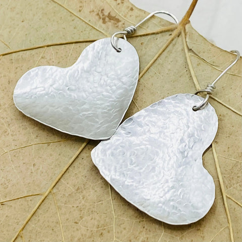JANECKA Sterling Silver Hand Forged Heart 1.5 Earrings by Janecka—two elegant heart-shaped earrings with hooks, beautifully presented on a dried, light brown veined leaf; a stunning handmade jewelry piece.