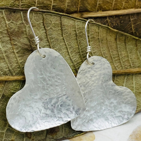 Janecka’s JANECKA Sterling Silver Hand Forged Heart 1.5 Earrings feature two heart-shaped sterling silver earrings with hooks displayed on a textured dried leaf background.