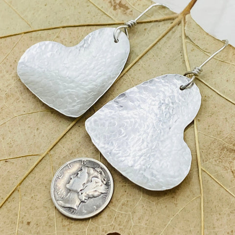 The Janecka Sterling Silver Hand Forged Heart 1.5 Earrings feature a textured finish and hook attachments, shown resting on a dried leaf next to a U.S. dime for size reference.
