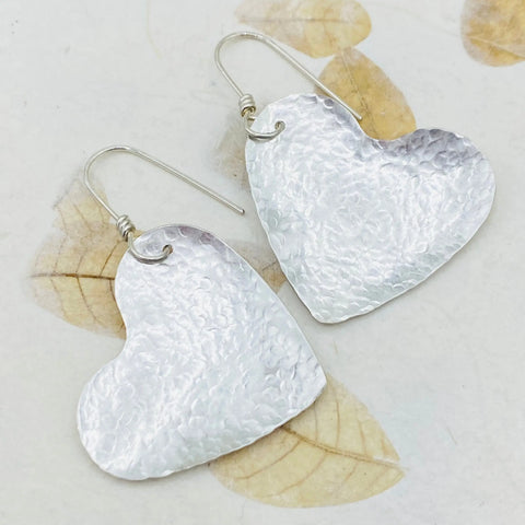 JANECKA Sterling Silver Hand Forged Heart 1.5 Earrings by Janecka—elegant, handmade heart-shaped earrings with hook clasps, shown on a light background with subtle dried leaf patterns.