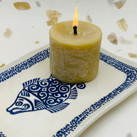A lit beige candle rests on the Janecka JANECKA Blue Fish Tray, a handmade pottery plate with blue fish design and border. Light brown leaves adorn the background, making it an ideal artisan gift.