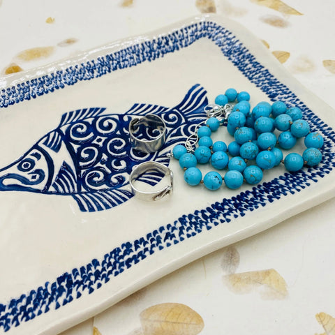 The JANECKA Blue Fish Tray by Janecka, crafted from handmade pottery, displays a turquoise beaded bracelet and two silver rings. This artisan gift sits on a white surface with subtle beige leaf patterns.