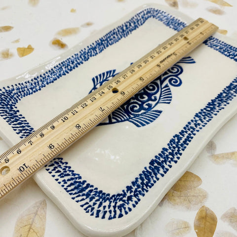 The JANECKA Blue Fish Tray by Janecka features a handmade rectangular design with a blue fish motif and patterned border, shown with a wooden ruler amid yellow leaves—a unique artisan gift for handcrafted pottery lovers.