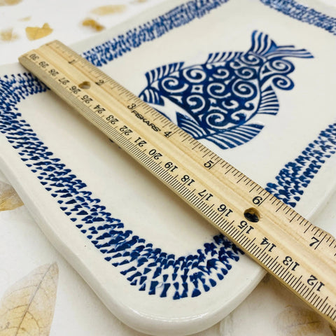 A wooden ruler measures the Janecka Blue Fish Tray, Handmade Pottery—a rectangular ceramic tray with a blue stylized fish and dotted border. The plate rests on a leafy white surface, making it an ideal artisan gift.