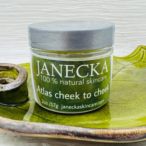A 2oz/57g jar of Janecka Skincare’s Atlas Cheek to Cheek Herbal Healing Ointment, made with natural ingredients, rests on a green ceramic dish. The label lists janeckaskincare.com.
