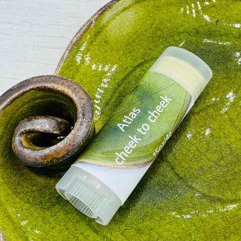 A tube labeled JANECKA Atlas Cheek to Cheek, Herbal Healing Ointment by Janecka Skincare, rests on a textured green ceramic dish with a curled accent. The product is an herbal cosmetic or skincare ointment.